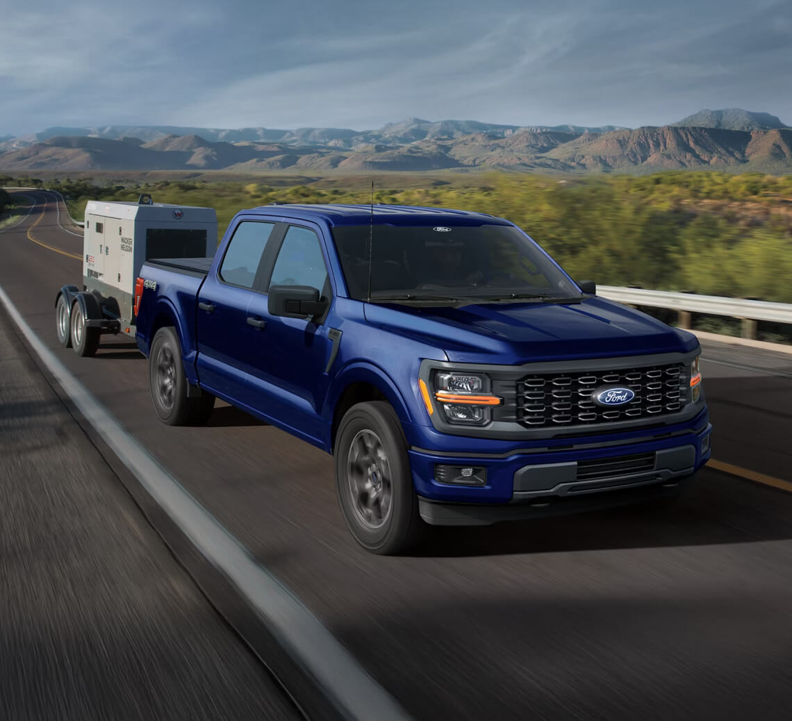 Front exterior action shot of a blue 2026 Ford F-150 towing a generator down a remote highway.